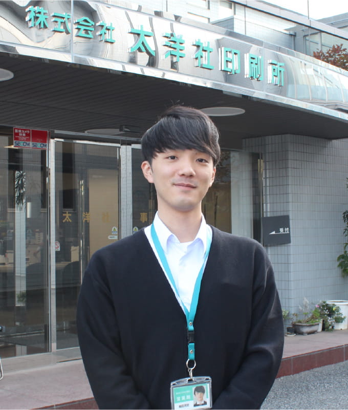 写真：梅田 河空さんのポートレート。こちらを向いて笑顔。背景には株式会社大洋社印刷所のエントランス