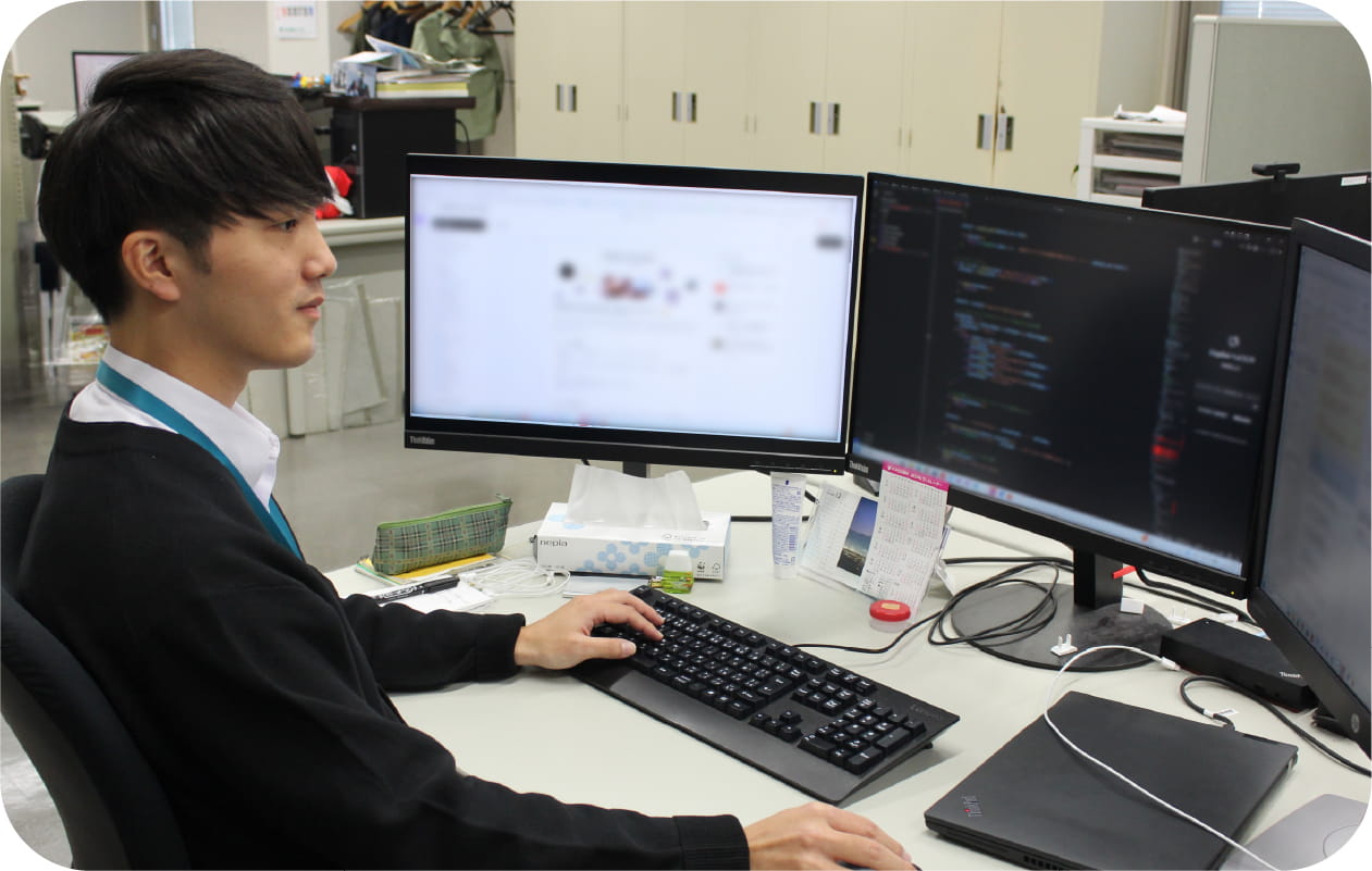 写真：3枚のディスプレイに向かってパソコンで作業をしている梅田さん